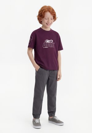 Vörös hajú fiú lila mintás pólóban, sötétszürke futó nadrágban és szürke fűzős sportcipőben, egyik kezét a zsebében tartva áll.