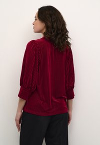 Blusa roja con plisados, confeccionada en un material de terciopelo elástico, con mangas abullonadas y un escote redondo, combinada con pantalones negros.