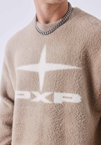 Suéter beige y suave con una gran estrella blanca y el logo "PXP". Diseño de cuello redondo con una textura suave y un collar de cadena plateada.