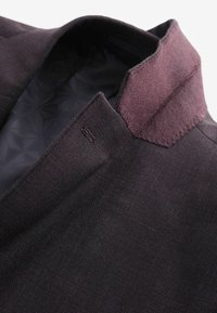 Mørk lilla blazer med en struktureret stof og en kontrastfarvet filtlignende krave. Indvendigt har den et geometrisk mønster, der forstærker den visuelle dybde.