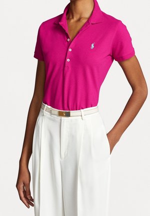 Kobieta ubrana w jasnoróżową koszulkę polo z krótkim rękawem, włożoną w wysokie białe spodnie z dopasowanym paskiem.