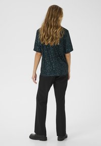 Zwart pailletten top met korte mouwen en een relaxte pasvorm, gecombineerd met zwarte pantalons en stevige zwarte schoenen, gezien van achteren.