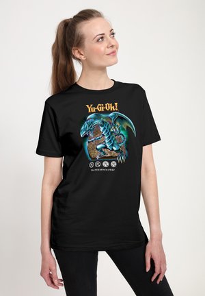 Ung kvinde iført en sort Yu-Gi-Oh! t-shirt med et blåt dragegrafik og japansk tekst, der kigger op og smiler let.