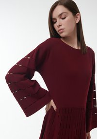 Robe tricotée bordeaux avec des manches larges dotées de découpes horizontales et un corsage texturé. Des fentes verticales ornent la partie inférieure pour un détail supplémentaire.