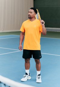 Orange short-sleeve athletic shirt, black shorts, white socks, and tennis shoes. Holding a tennis racket. Indoor court setting.