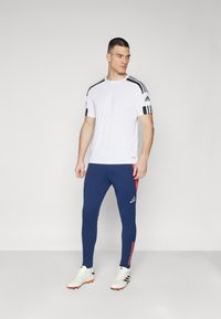 Hvid atletisk t-shirt med sorte striber på skulderne, parret med blå leggings med røde sideaccenter, båret med hvide fodboldstøvler.