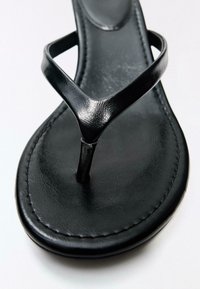 Zwarte lederen teensandaal met een gestikte zool en een glanzende V-vormige band over het voetbed op een witte achtergrond.