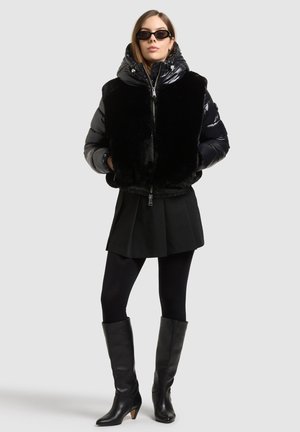 Un mannequin pose devant un fond uni, portant des lunettes de soleil, une doudoune noire bordée de fourrure, une jupe, un legging et des bottes montantes jusqu’aux genoux.