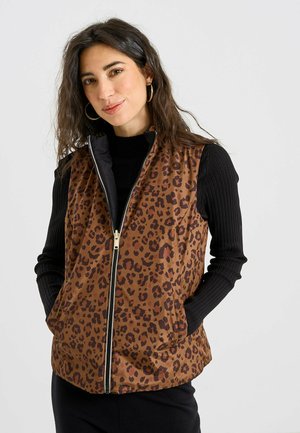 Mujer con un chaleco marrón sin mangas con estampado de leopardo y cremallera, encima de una camiseta negra de manga larga con canalé, de pie con las manos en los bolsillos.