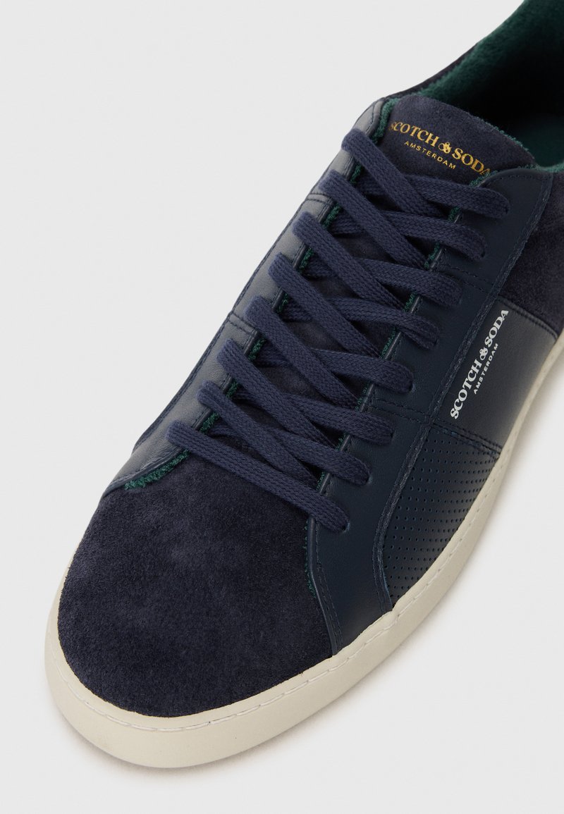 Baskets en daim et cuir bleu marine avec une finition lisse, dotées de lacets foncés, d'un panneau perforé et d'une semelle en caoutchouc blanche. Détail du logo doré.