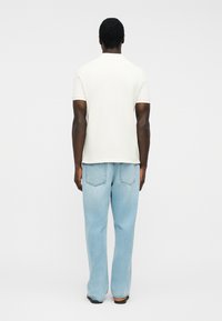 Hombre de pie, de espaldas, vistiendo una camisa blanca de manga corta sencilla y vaqueros azul claro de corte holgado, frente a un fondo liso.