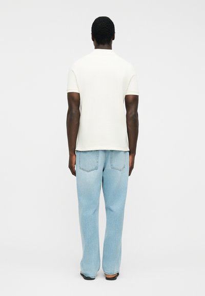 Homme debout de dos, portant un t-shirt blanc uni à manches courtes et un jean bleu clair ample, sur un fond uni.