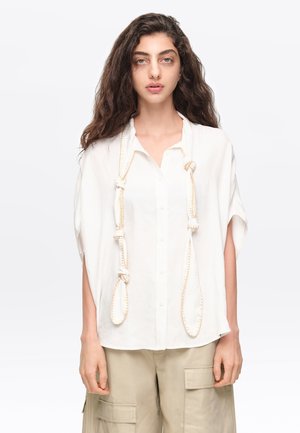 Mujer con cabello largo y rizado que lleva una camisa blanca holgada con botones y detalles de cuerda anudada, y pantalones cargo beige, de pie frente a un fondo blanco.