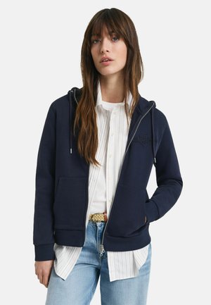 Granatowy hoodie z zamkiem wykonany z miękkiego materiału, z detalem logo z przodu, dwiema bocznymi kieszeniami i ściągaczami przy mankietach, nałożony na białą koszulę w paski.