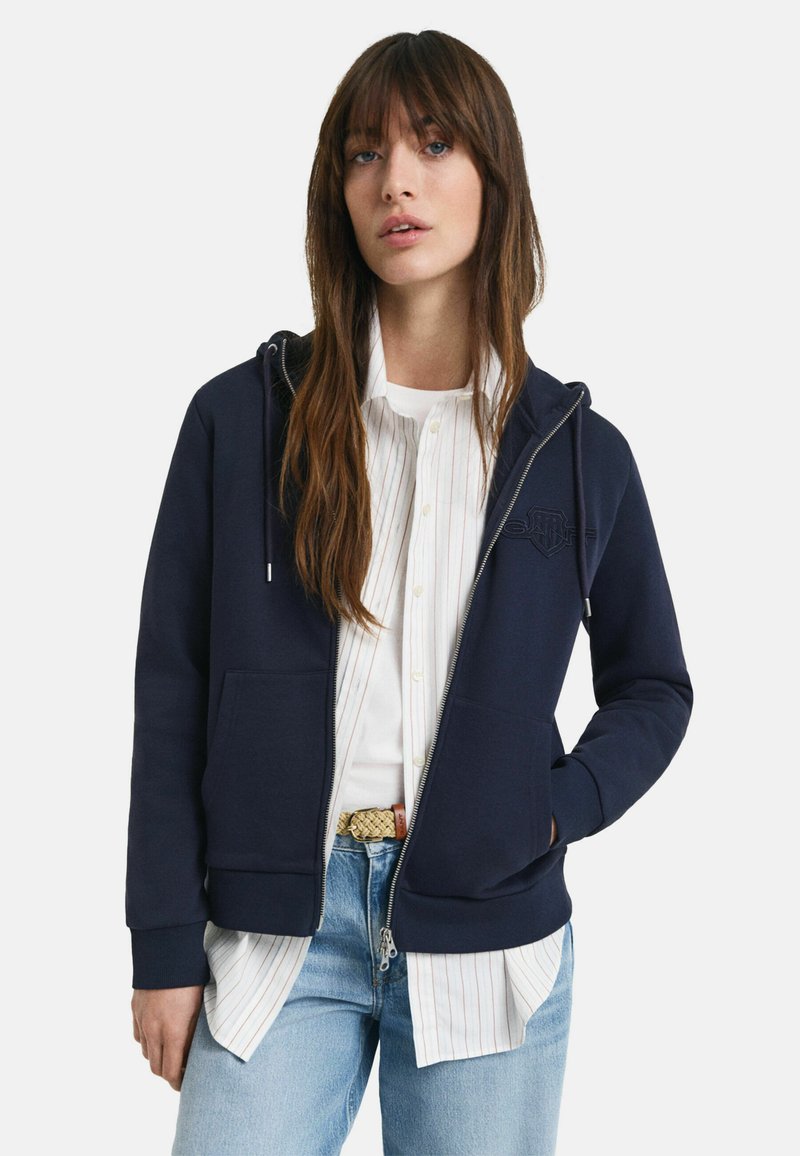 Sudadera con cremallera en color azul marino, fabricada en un material suave, que presenta un detalle de logo en la parte delantera, dos bolsillos laterales y puños acanalados, llevada sobre una camisa blanca a rayas.