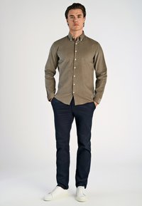 Olijfgroene shirt met een knoopsluiting, lange mouwen en witte knopen, gecombineerd met marineblauwe broeken en witte sneakers.