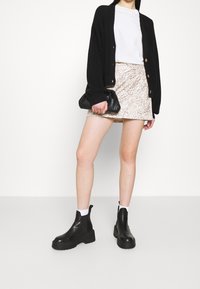 Kvinna iklädd svart kofta, vit t-shirt, ljus mönstrad minikjol, svarta ankelboots, hållande en liten svart clutch-väska.