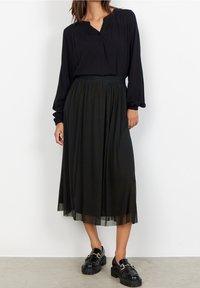 Frau in einer schwarzen Bluse mit langen Ärmeln, einem schwarzen midi Faltenrock und schwarzen Slippern mit goldenen Kettendetails, steht vor einer weißen Wand.
