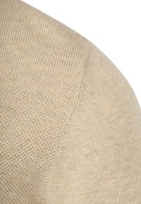 Beige Strickpulloverstoff mit dichter Webart und subtiler Textur, mit einer Naht an der Schulter und leichtem Glanz.