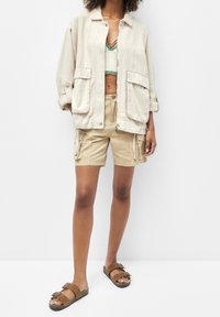 Veste en coton beige clair avec deux poches avant, associée à un short kaki et des sandales brunes à glissière. Accent vert sur le crop top.