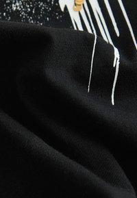 Tela de algodón negro con una textura suave, que presenta detalles gráficos en blanco y acentos dorados. El material parece suave y ligeramente arrugado.