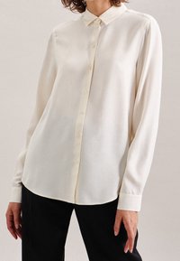 Woman wearing a long-sleeve cream button-up blouse paired with black trousers against a plain background.