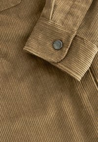 Tissu en velours côtelé marron avec une manche retroussée fermée par un bouton noir rond gravé de texte autour du bord.