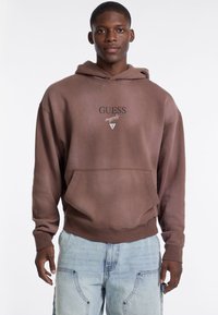 Felpa con cappuccio marrone, dotata di tasca frontale, con logo "GUESS" nero e grafica triangolare. Maniche e orlo a costine. Vestibilità casual, tessuto morbido.
