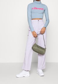 Hellblaue, kurze Rollkragen-Bluse mit pinkem "ellesse"-Logo, kombiniert mit lavendelfarbenen Cargo-Hosen und einer olivgrünen Umhängetasche. Weiße Sneakers sind ebenfalls enthalten.