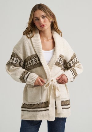 Jonge vrouw die een riem vastmaakt aan een crèmekleurige gebreide cardigan met bruine gestreepte patronen en voorzakken, draagt een witte top en blauwe spijkerbroek.