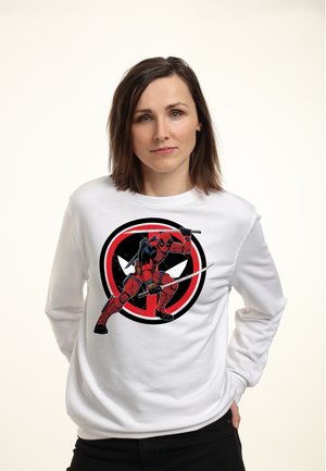 Vrouw met schouderlang bruin haar die een witte trui draagt met Deadpool die zwaarden vasthoudt voor een rood-zwart logo.