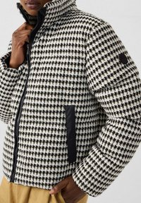 Persoon in een zwart-wit houndstooth-patroon pufferjack met zwarte rits en zakdetails, hand die de kraag vasthoudt.