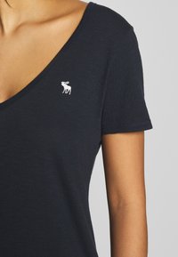 Tmavě modré bavlněné tričko s hlubokým výstřihem do V a krátkými rukávy. Obsahuje malé bílé vyšívané logo losa na hrudi.