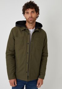 Grön bomulljacka med svart huva, med två bröstfickor, knäppning med knappar och en strukturerad design. Bärs över en vit skjorta.