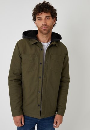 Zielona bawełniana kurtka z czarnym kapturem, wyposażona w dwie kieszenie na piersi, z zapięciem na guziki oraz o stonowanej konstrukcji. Noszona na białej koszuli.