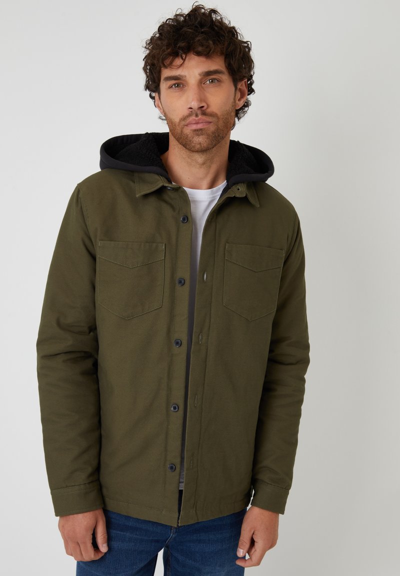 Grön bomulljacka med svart huva, med två bröstfickor, knäppning med knappar och en strukturerad design. Bärs över en vit skjorta.