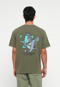 Olivengrøn bomulds T-shirt med et stort grafisk print af en flyvende and, "TJ" initialer i turkis og teksten "TOMMY JEANS New York City".