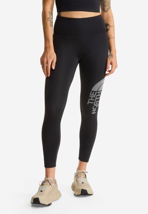 Vrouw draagt zwarte leggings met hoge taille met groot "The North Face" logo op dij en beige sportschoenen, staand tegen een witte achtergrond.
