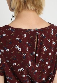 Burgunderfarbener Blumentop mit weißen und blauen Blumen, Knopfverschluss und einem kleinen Schlitz auf der Rückseite. Leichter Stoff mit Struktur.