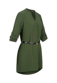 Tunique verte à manches longues avec manches retroussées et ceinture noire avec boucle double anneau dorée autour de la taille sur fond blanc.