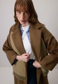 Vrouw met lichtbruin haar die een bordeauxbruine jas met twee tinten draagt over een blauw gestreept shirt en een witte top, poseert met haar handen terwijl ze de jas aanpast.