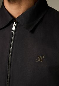 Schwarze Jacke mit Kragen, ausgestattet mit einem silbernen Reißverschluss und einem dezenten, goldenen, gestickten Logo auf der Brust. Glatte Textur, minimalistisches Design.