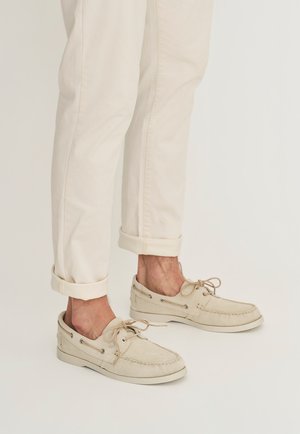 Benedenbenen met opgerolde crèmekleurige broeken en beige suède bootsschoenen met veters, staand op een effen lichte achtergrond.