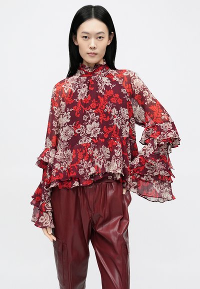 Áttetsző, piros virágos blúz fodros ujjakkal, magas nyakkal és réteges fodrokkal; magas derekú, bordó bőr nadrággal párosítva.