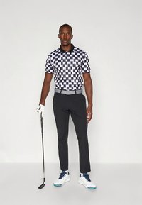 Polo de manga curta aos quadrados em preto e branco com gola preta, combinado com calças pretas e sapatos de golfe brancos. Segurando um taco de golfe.
