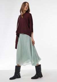 Bordeaux oversized trui, vloeiende mintgroene chiffonrok en zwarte kniehoge laarzen met gespaccenten, wat samen een gelaagde look creëert.