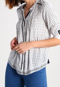 Blusa a maniche triple con motivo bianco e nero, dotata di un'apertura con bottoni, pieghe e orlo arrotondato; realizzata in tessuto leggero.