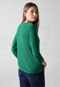 Maglione verde strutturato con scollo rotondo e maniche a tre quarti, abbinato a pantaloni in denim scuro, vista da dietro.