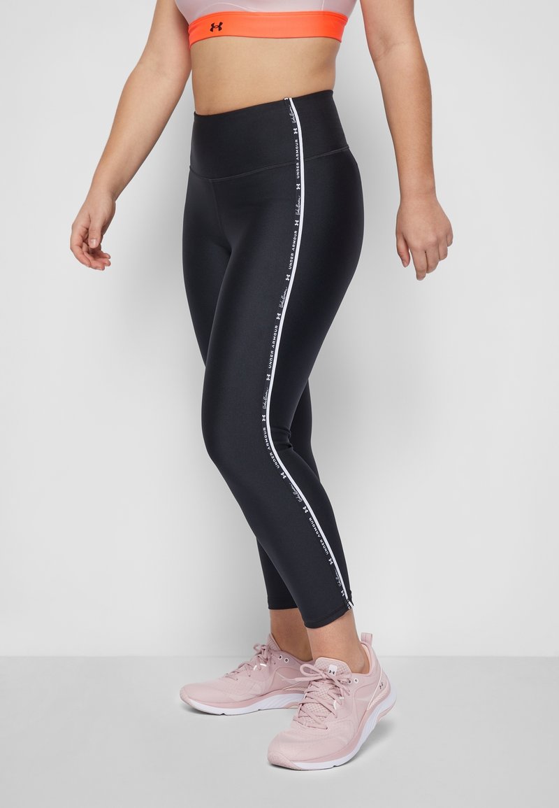 Leggings neri con vita alta presentano strisce laterali bianche e testo brandizzato. Il modello indossa scarpe da ginnastica rosa chiaro.