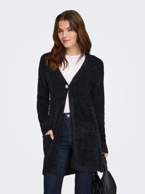 Černý, chlupatý svetr s otevřeným předním dílem, dlouhými rukávy, zapínáním na jeden knoflík a dvěma bočními kapsami, nošený přes bílý top a džíny.